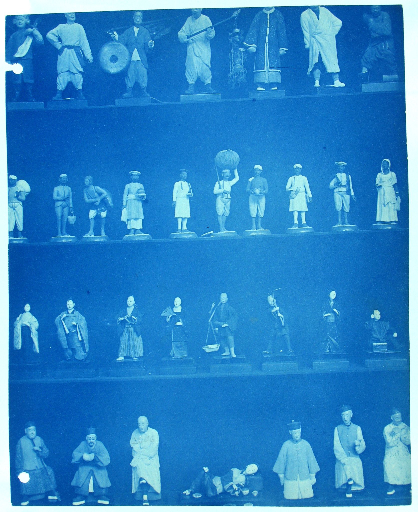 A vintage photograph of a collection of small figurines or statues displayed on shelves. The photograph has a strong blue tint, giving it a historical and somewhat ethereal quality.
