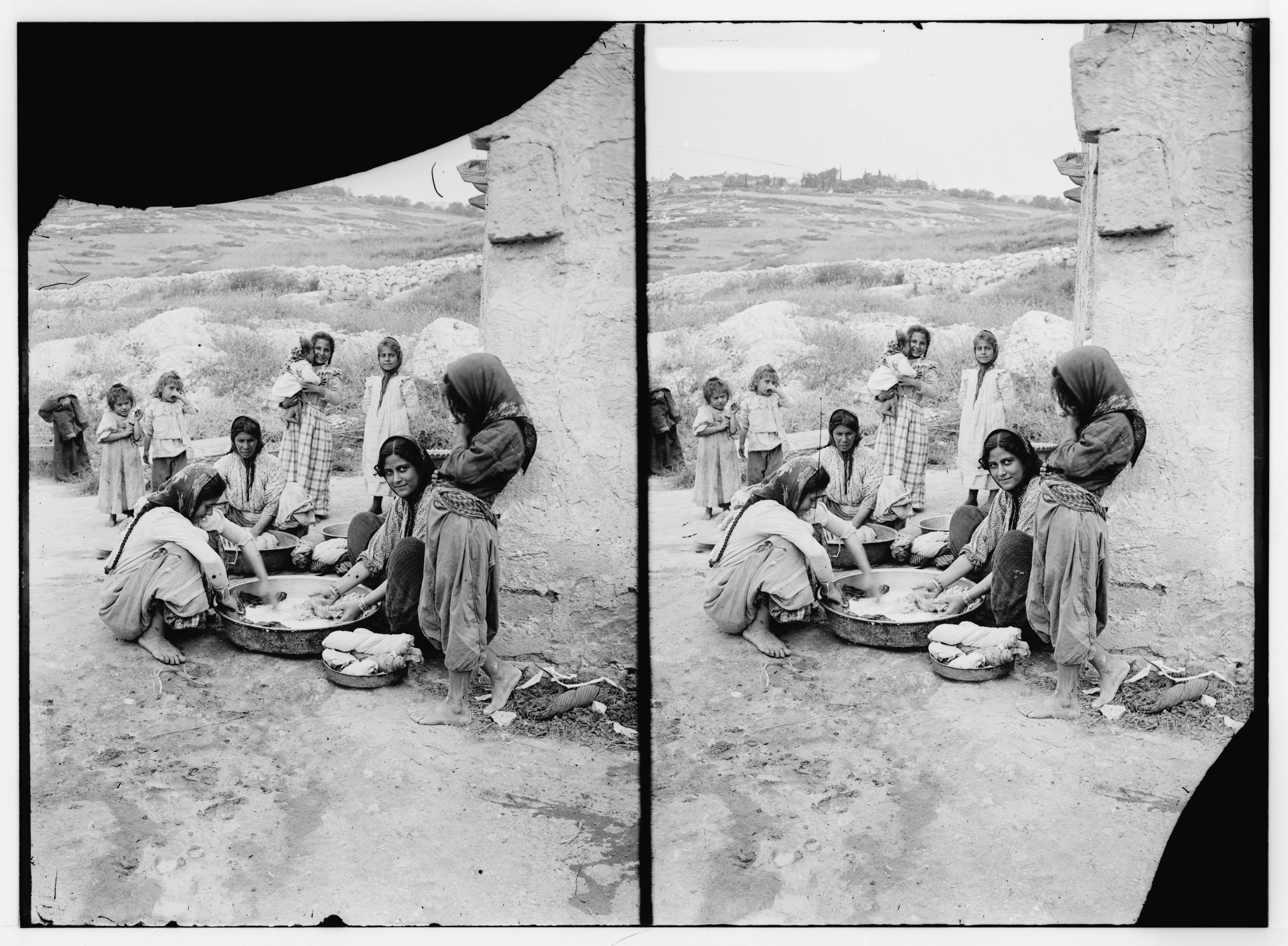 Stereoscopic photograph of a group of people gathered outdoors, likely a family or community, engaged in a communal activity. Women are seated around bowls, and children are standing and watching.