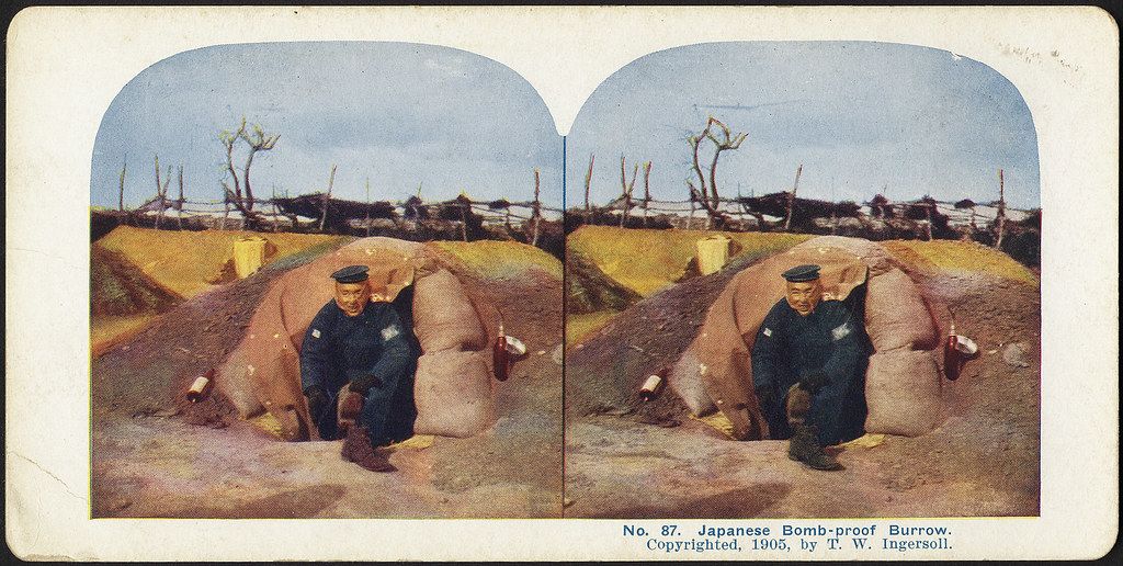 Stereoscopic photograph of a Japanese soldier standing in front of a bomb-proof burrow constructed from sandbags, 1905.