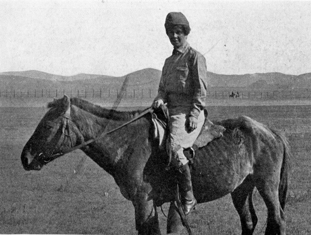Vintage black and white photo of a young man in uniform riding a horse across a grassy plain with rolling hills in the background.