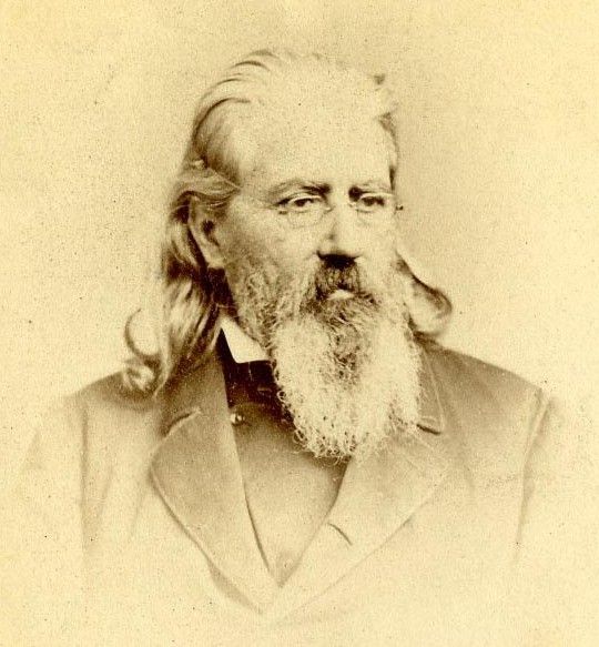 Sepia-toned portrait of an older man with a long white beard and hair, wearing a dark suit.