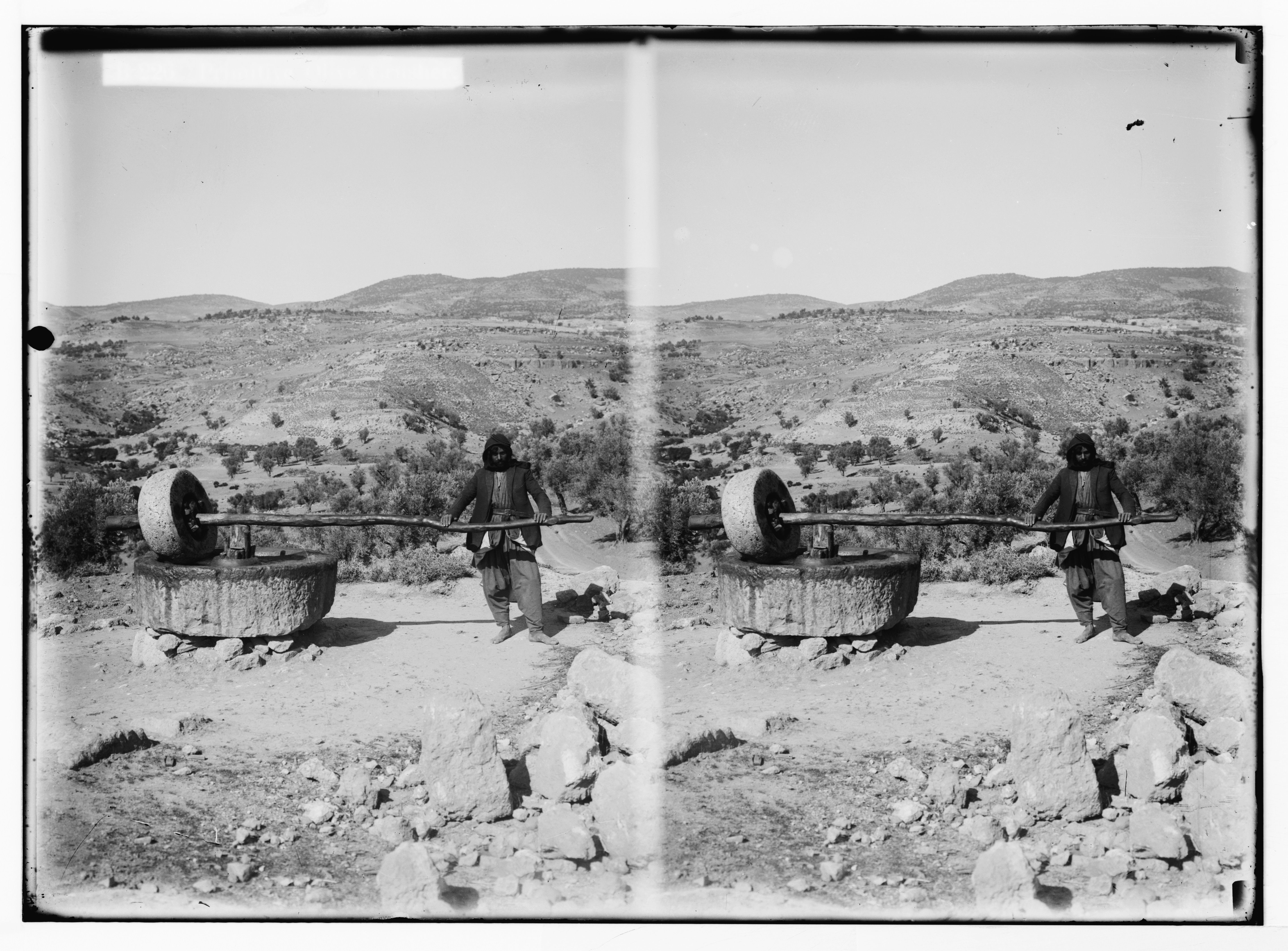 Stereoscopic photograph of a person standing next to a stone structure with a wheel in a hilly landscape.