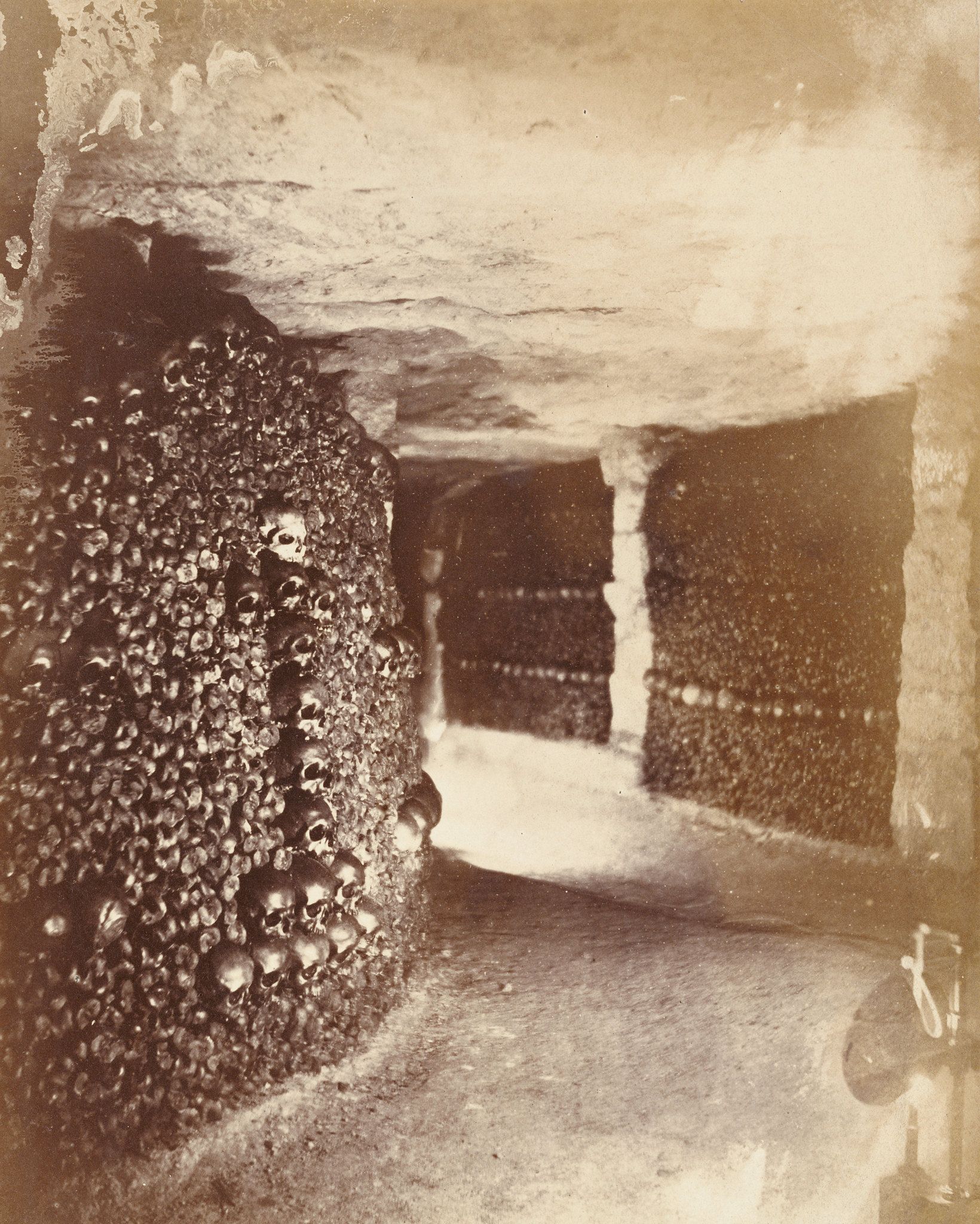 A sepia-toned vintage photograph of the Paris Catacombs, showing walls and arches constructed entirely of human skulls and bones.
