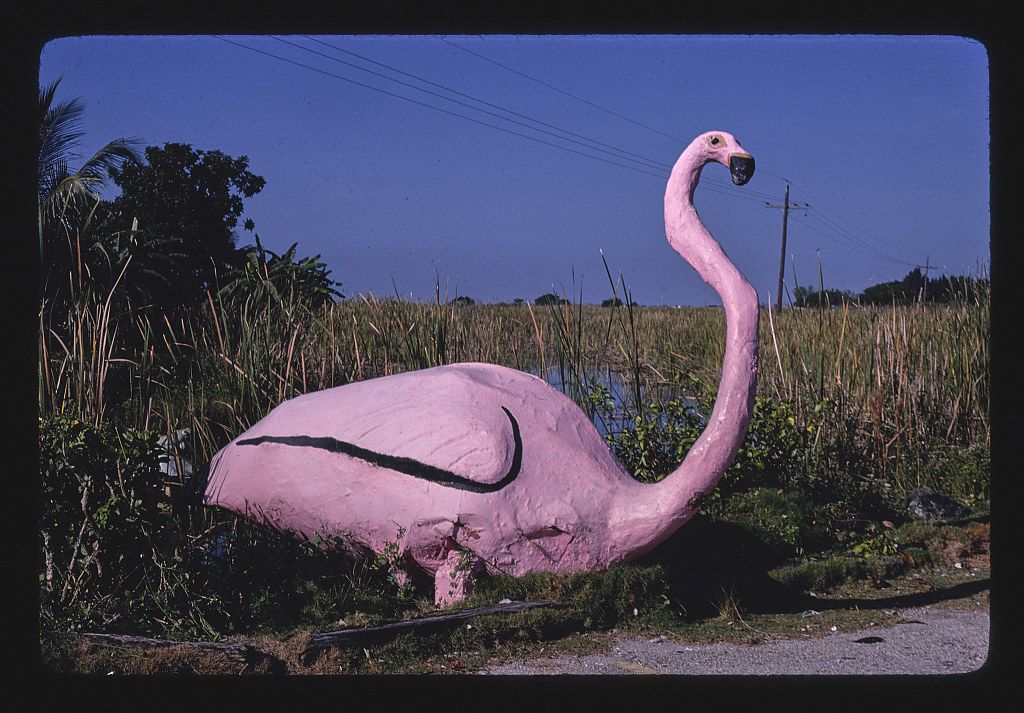 A large, pink flamingo statue stands in a field of tall grass near a road, with trees and a dark blue sky in the background.