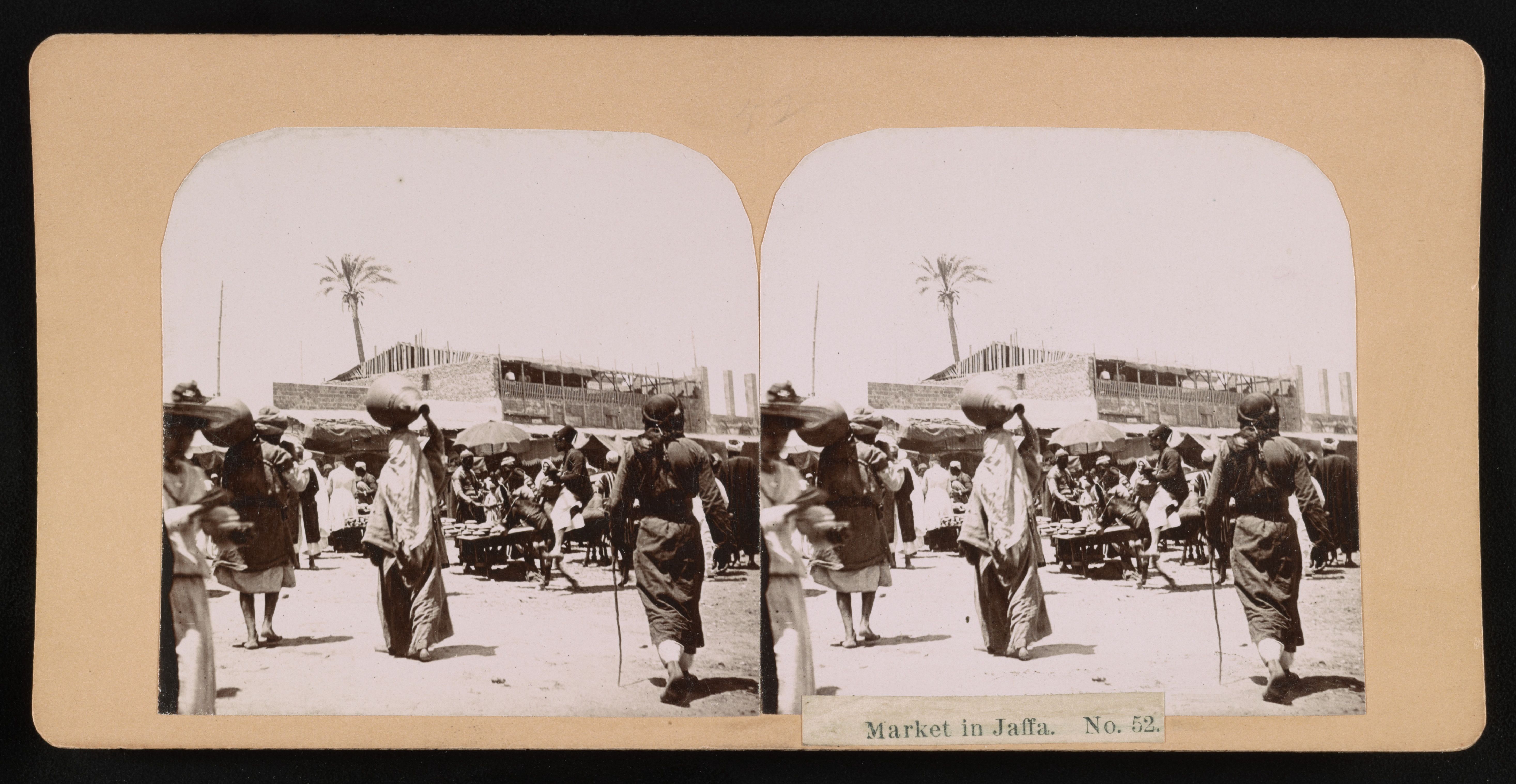 Stereoscope card of a market in Jaffa, featuring people in traditional clothing, women carrying baskets on their heads, and a bustling open-air market scene.