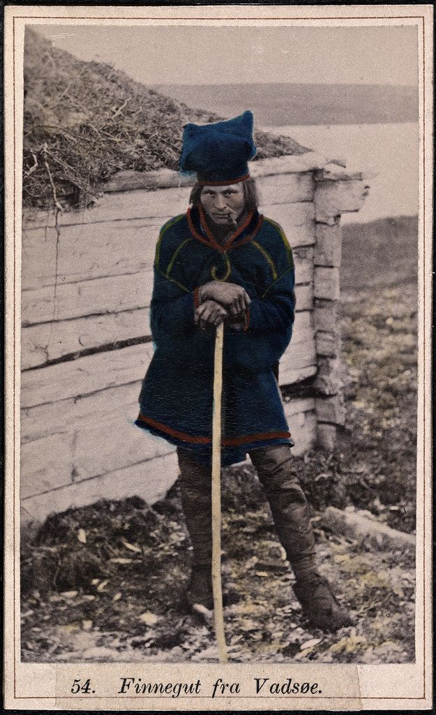 Vintage hand-colored photograph of a Sami man in traditional clothing standing outdoors.