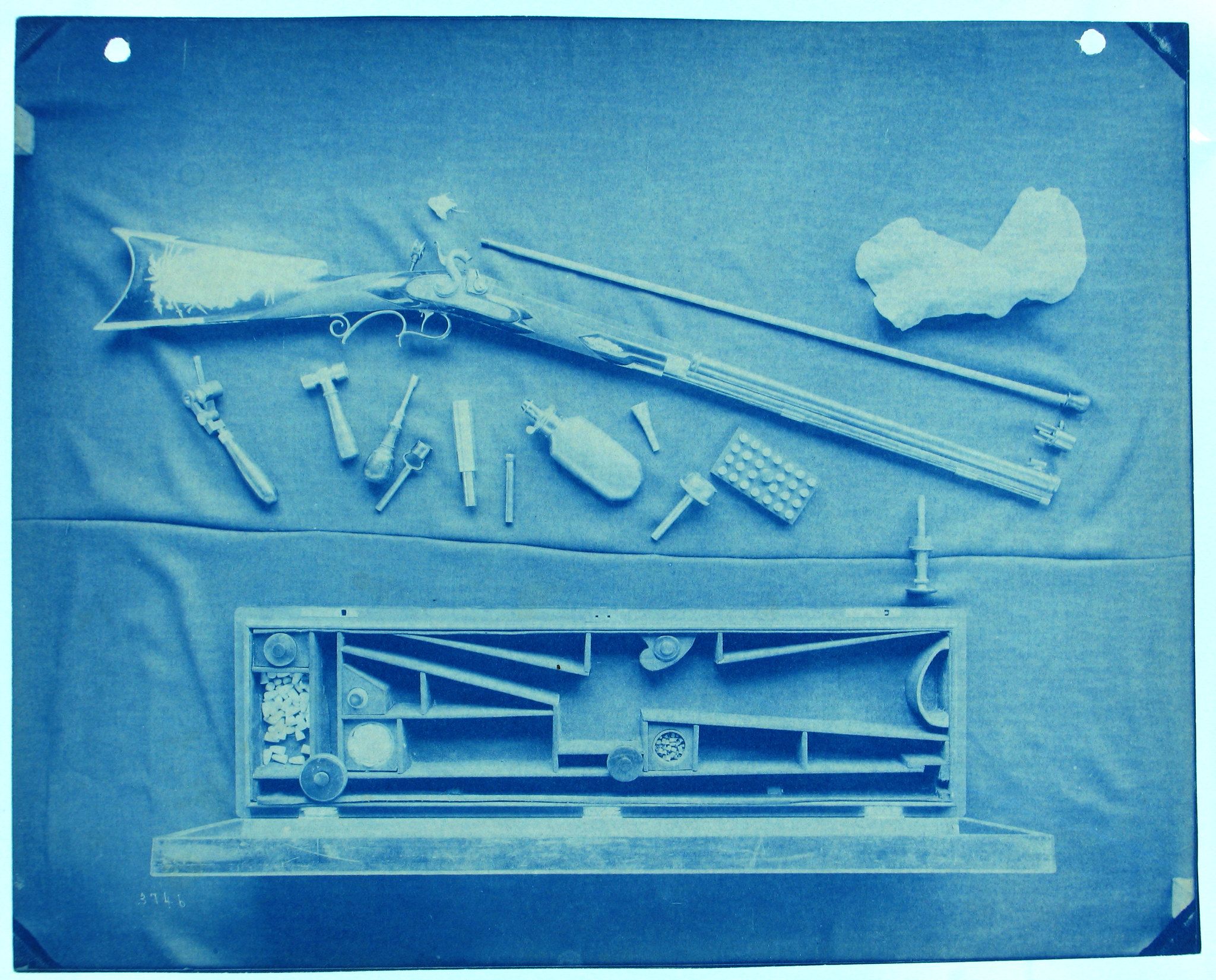 Cyanotype photograph of a gun, tools, and an open box on a blue cloth.