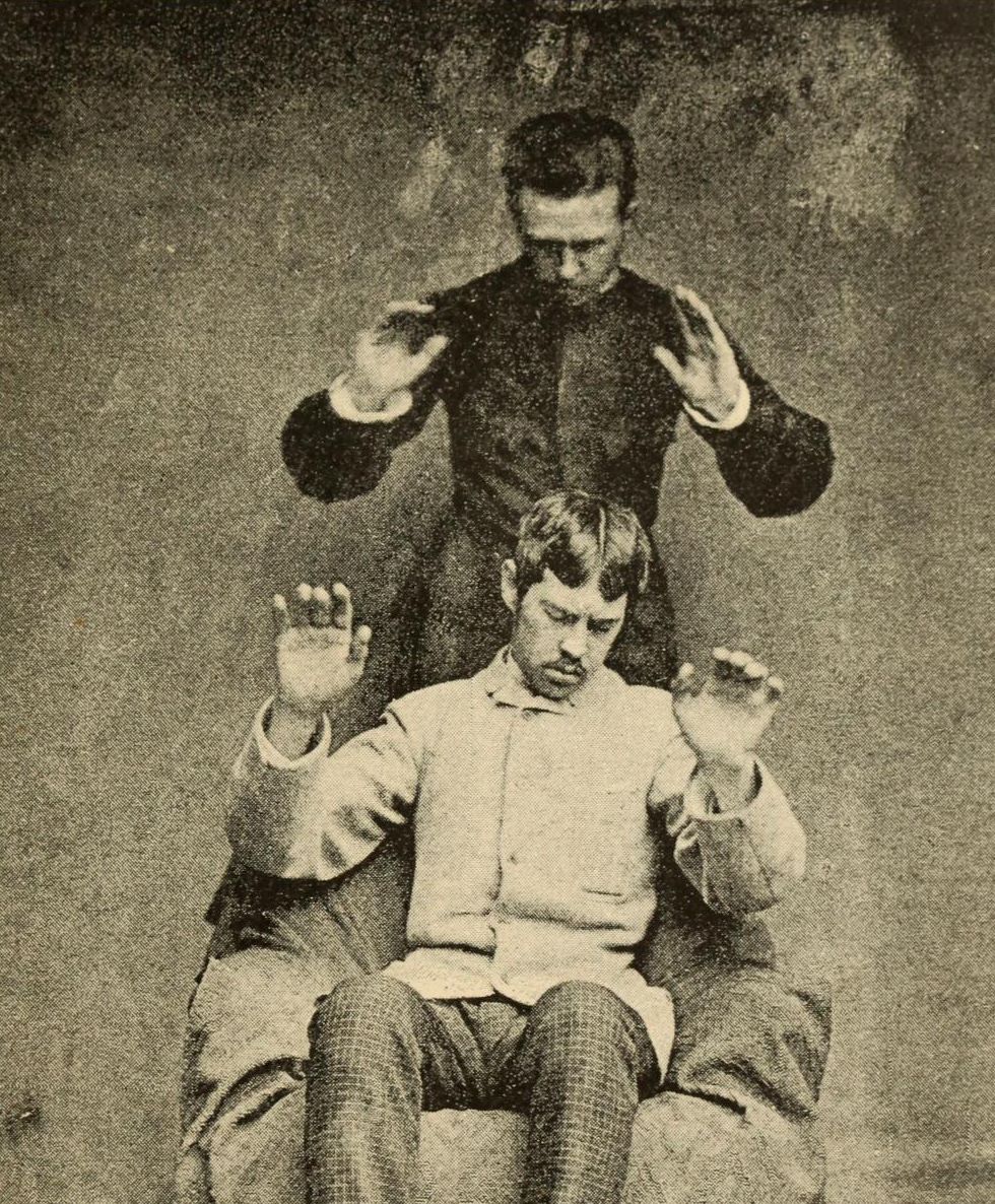 Vintage black and white photograph of two men in a staged pose, one seated and one standing behind, with raised hands.