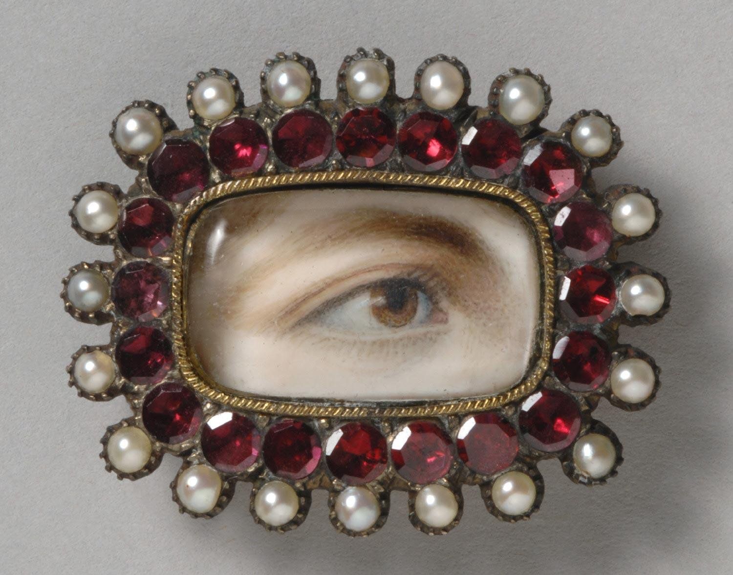 Close-up of a vintage brooch featuring a painted eye surrounded by red garnets and pearls.