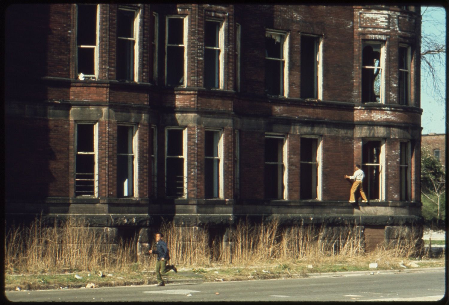 A large, brick building with numerous empty window frames. The building appears to be abandoned or severely damaged, with blackened brick suggesting a fire. The windows are mostly empty, with some showing remnants of frames or broken glass. In the foreground, a street runs parallel to the building. A person is seen running across the street, and another is standing near the base of the building. The area between the street and the building is overgrown with dry, golden-brown grass and scattered debris.