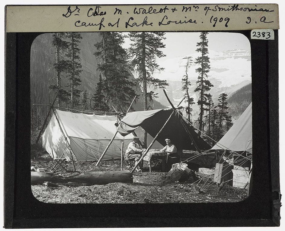 Black and white photograph of a campsite with tents, trees, and people in a forest setting, labeled as "Dr. Chas. M. Walcot & Mr. of Smithsonian Camp at Lake Louise. 1909".