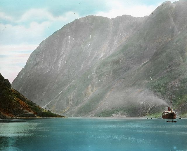Vintage photograph of a fjord with towering cliffs, a steamship, and a blue sky.