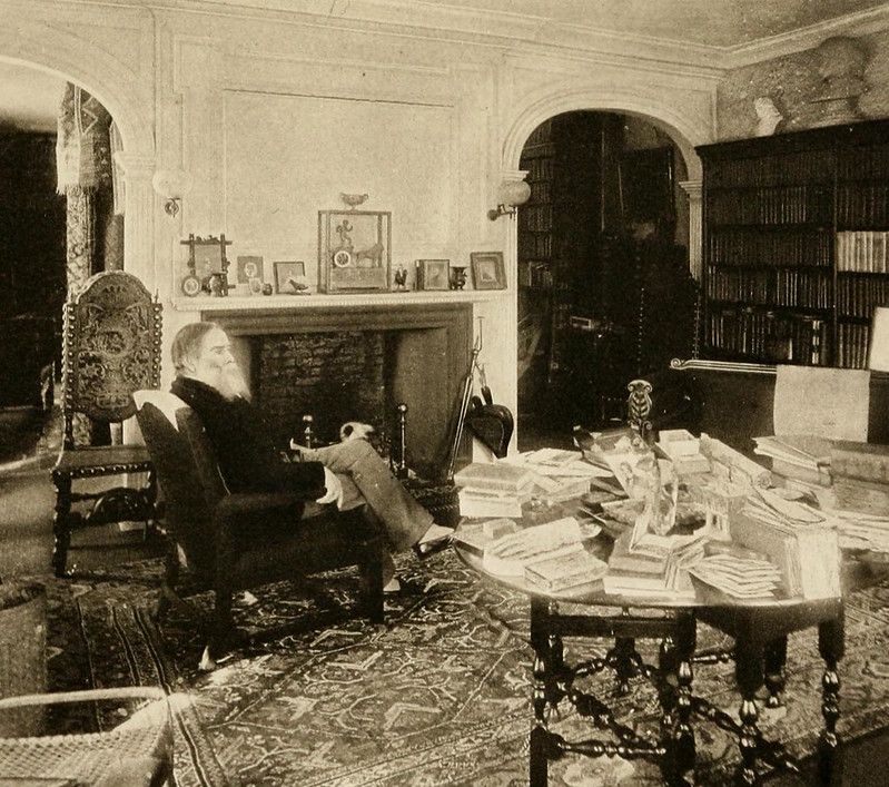 A black and white photograph of a person seated in a chair with a doll, surrounded by books and a fireplace in a richly decorated room.