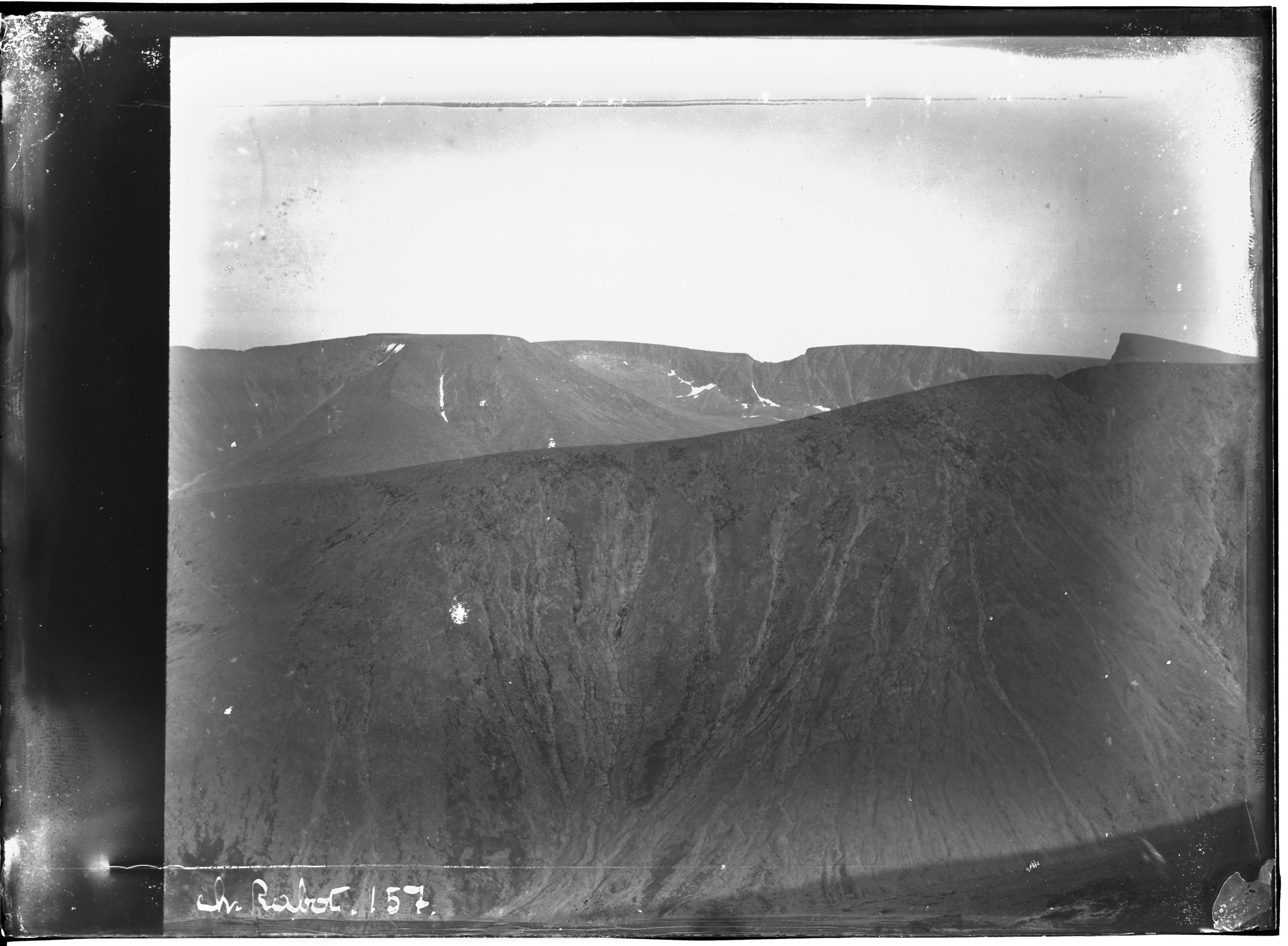 A vintage black and white photograph of a dramatic mountain landscape with a steep hillside in the foreground and a flat-topped mountain rising in the midground.