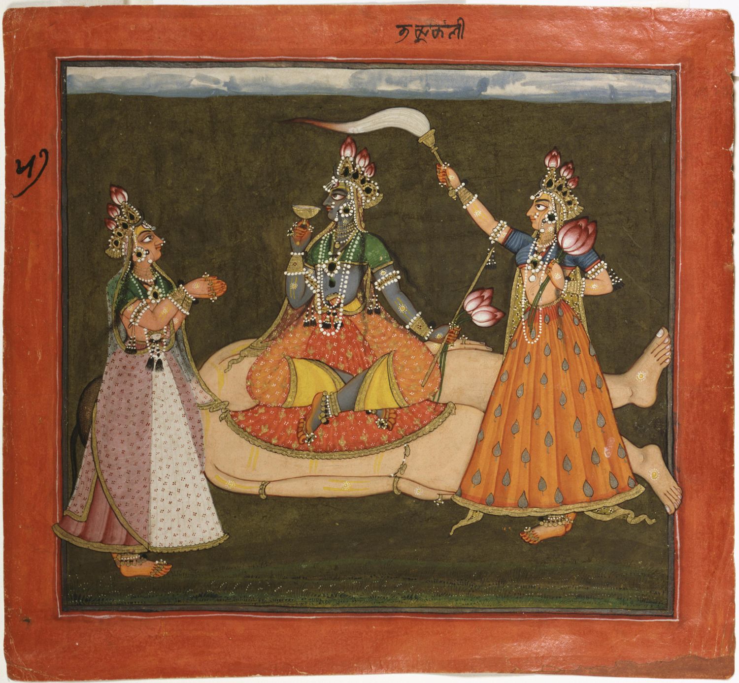 A painting depicting a scene with three figures. The central figure is a dark-skinned man reclining on a patterned orange and red cushion. He is adorned with a tall, ornate headdress, elaborate jewelry, and wears a dark blue garment. He holds a flute in his hands and appears to be playing it. On either side of him are two women, both dressed in flowing, patterned garments with orange teardrop-shaped motifs. They are also heavily adorned with jewelry, including necklaces, earrings, and armlets. One woman is playing a stringed instrument, while the other is holding a flaming object aloft. The background is a dark green, and the ground is covered with a patterned surface. The painting is framed by a red border. Above the central figure, there is a script in a language that is not immediately recognizable.