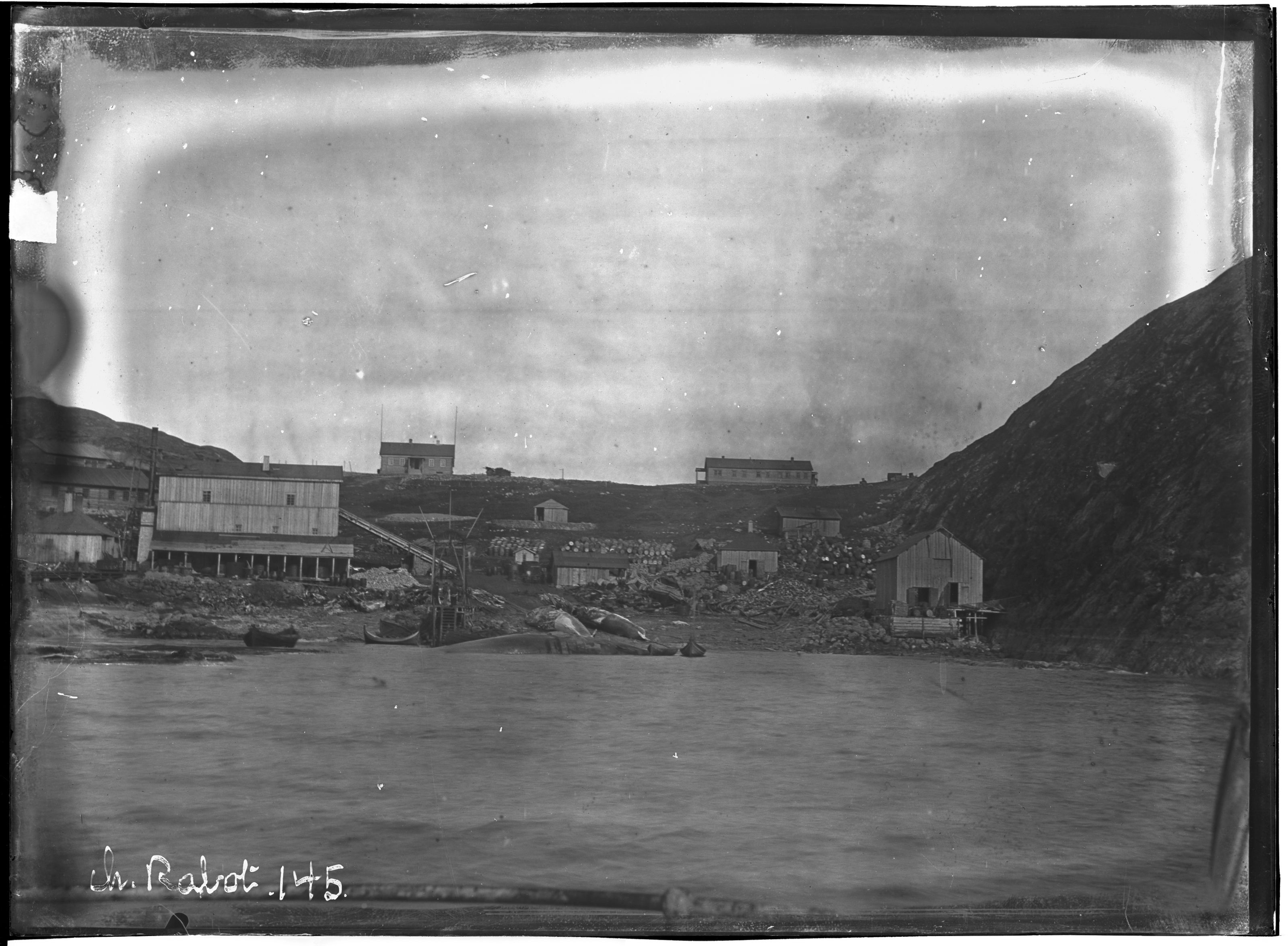 A vintage black and white photograph of a coastal settlement with wooden buildings, a dock, and a mountainous backdrop.