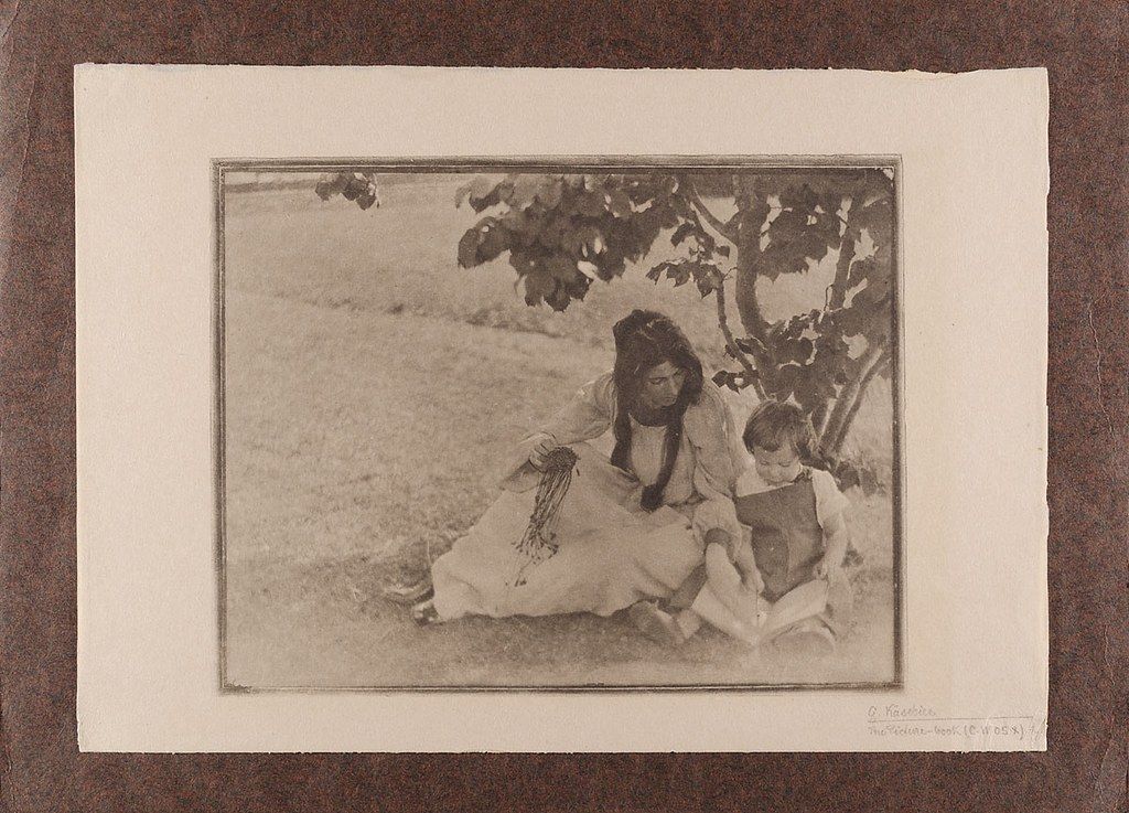 Vintage sepia-toned photograph of a Native American woman braiding a rope with a child reading a book beside her under a tree.