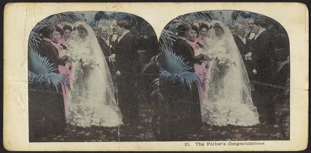 Stereoscope card titled "The Father's Congratulations" showing a bride in a decorated white dress being embraced by a man in a suit, with other guests in the background.