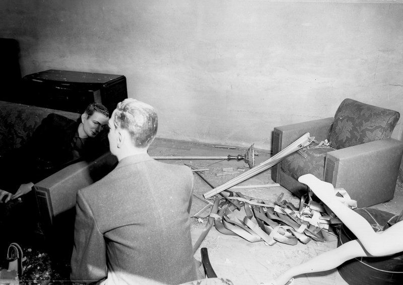 A black and white photograph of a man looking at a pile of debris in a room, with a broken piano and a second person visible in the background.
