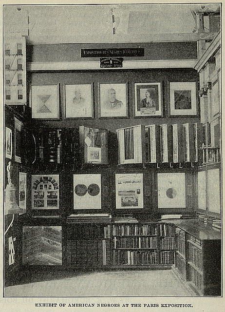 Black and white photograph of an exhibit titled "Exhibit of American Negroes at the Paris Exposition". The room is filled with displays, primarily consisting of framed pictures and books arranged on the walls.
