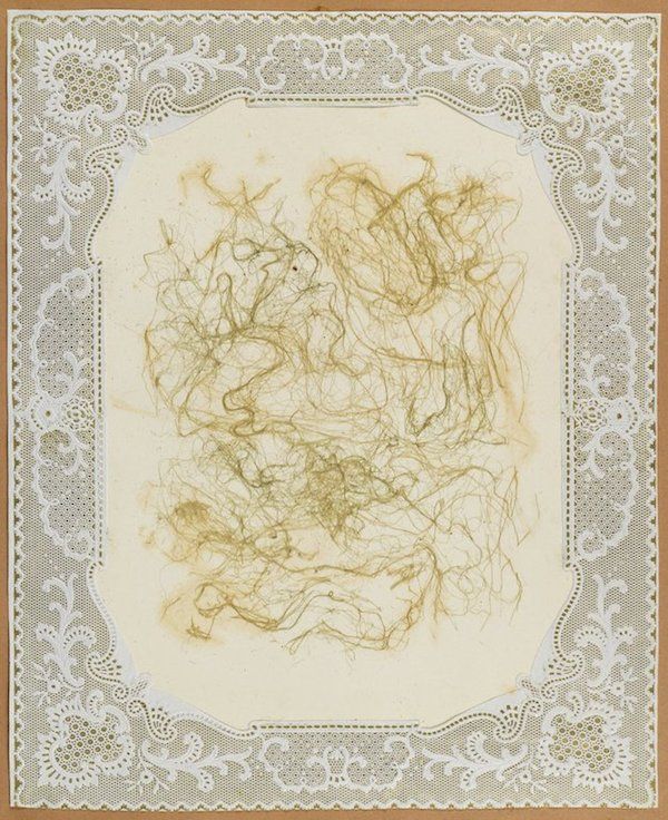 A vintage-style composition featuring a cluster of golden-brown hair arranged on cream-colored paper, framed by an ornate white lace border.