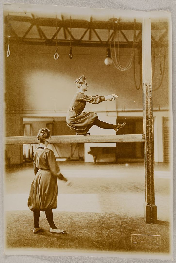 A vintage sepia-toned photograph of two young women in a gymnasium, one balancing on a beam and the other offering guidance.