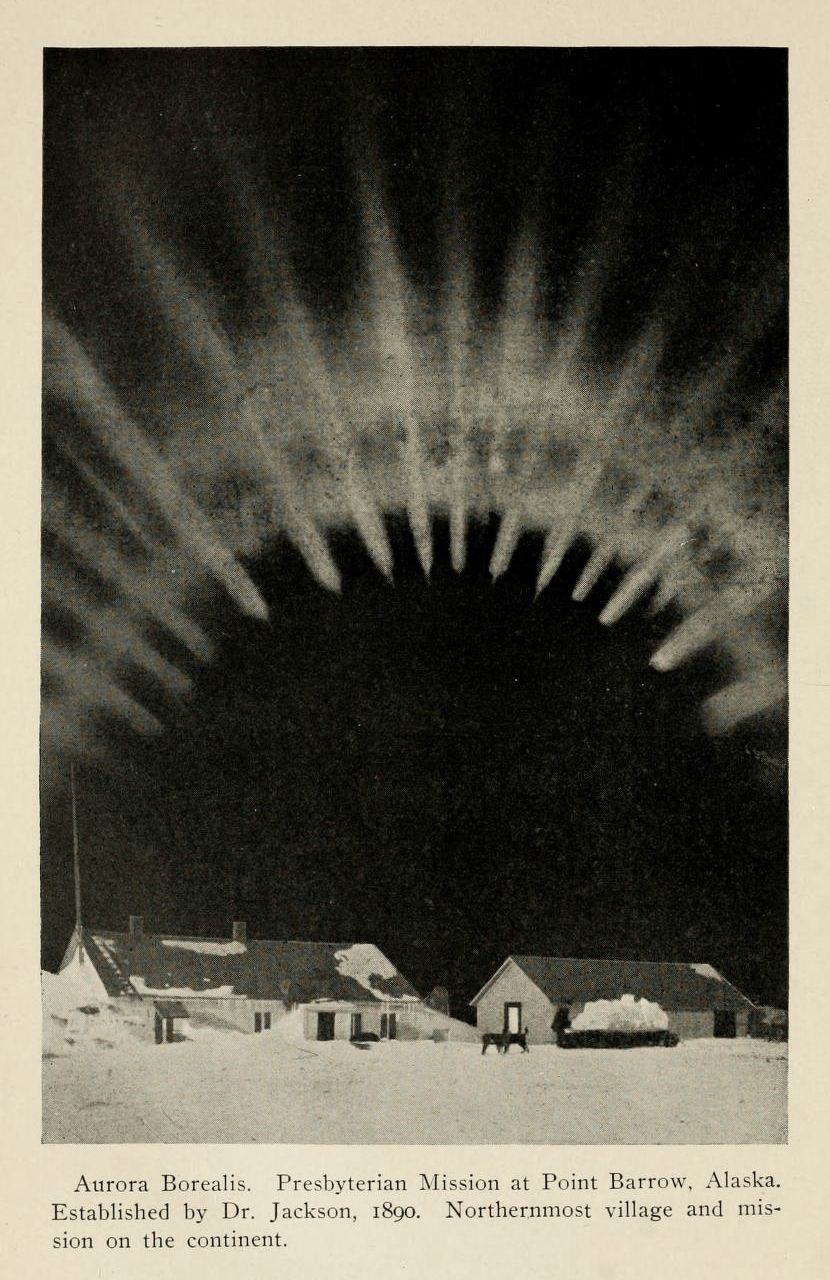 A vintage black and white photograph of the Aurora Borealis over a small village at Point Barrow, Alaska.