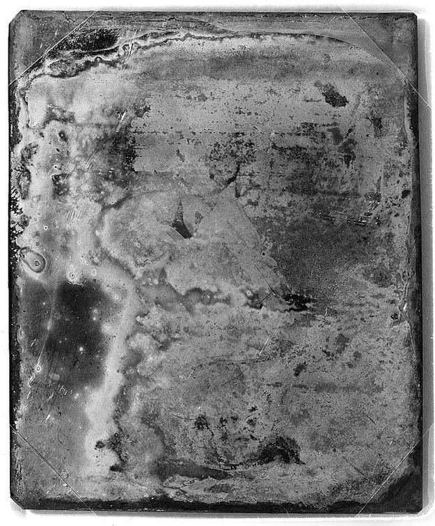 A daguerreotype plate with a blurred and faded image, showing a subtle pattern of light and dark areas, scratches, and blemishes.