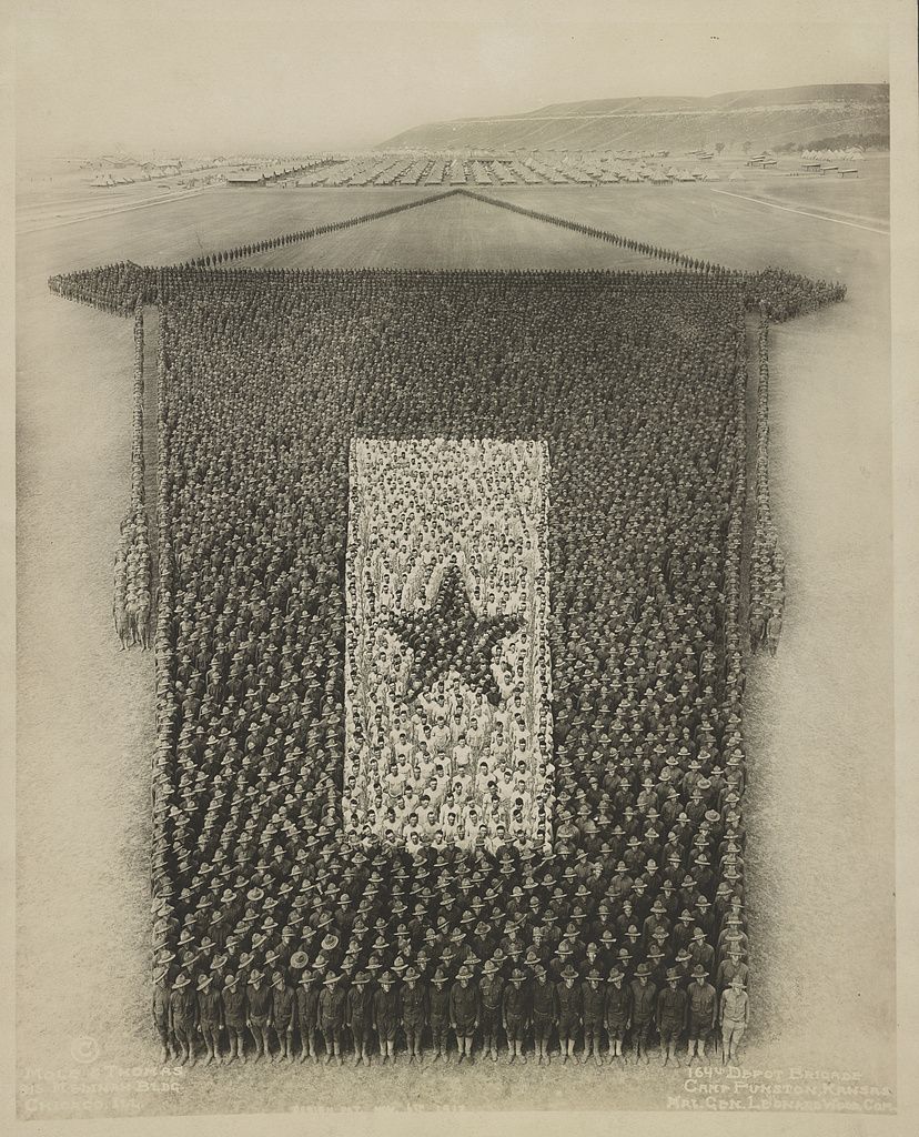 A vintage black and white photograph of a massive formation of soldiers in a field, forming a large rectangle with a lighter central space.