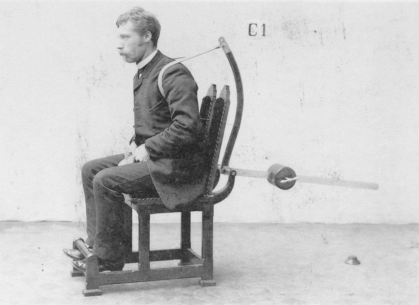 A black and white photograph of a man seated in a chair-like device with restraints.