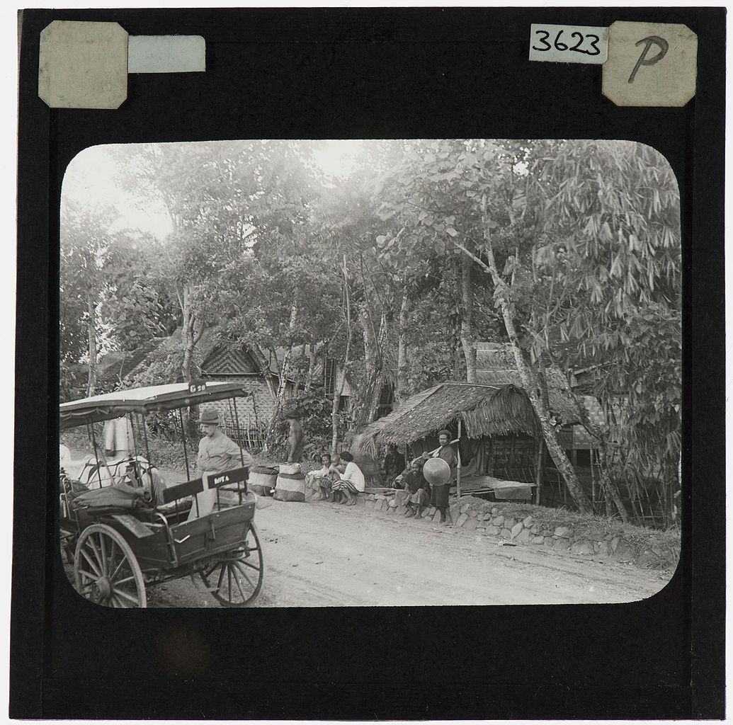 Black and white photograph of a vintage vehicle parked on a dirt road in a rural setting, with people gathered nearby and simple huts surrounded by trees.