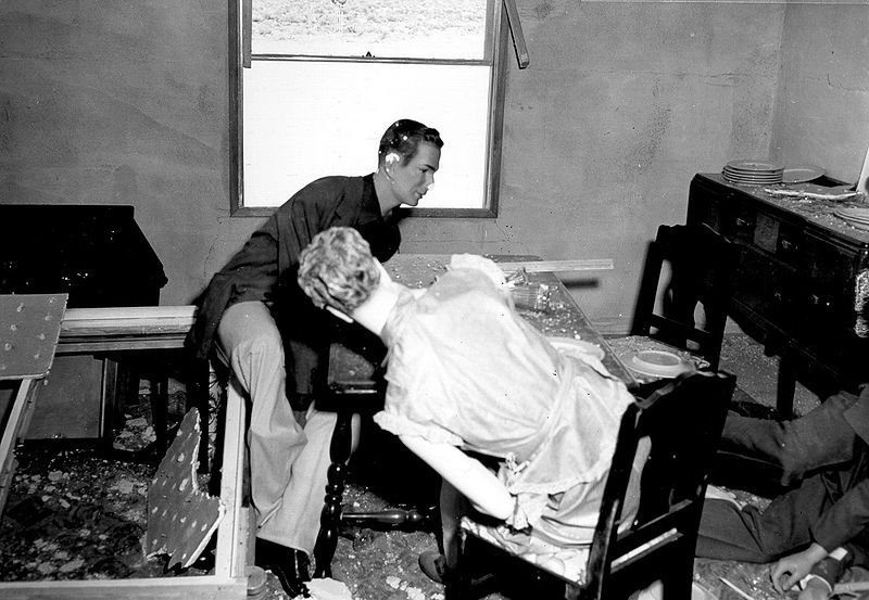A black and white photograph of a man checking on a woman lying in a damaged bed, surrounded by debris in a room.
