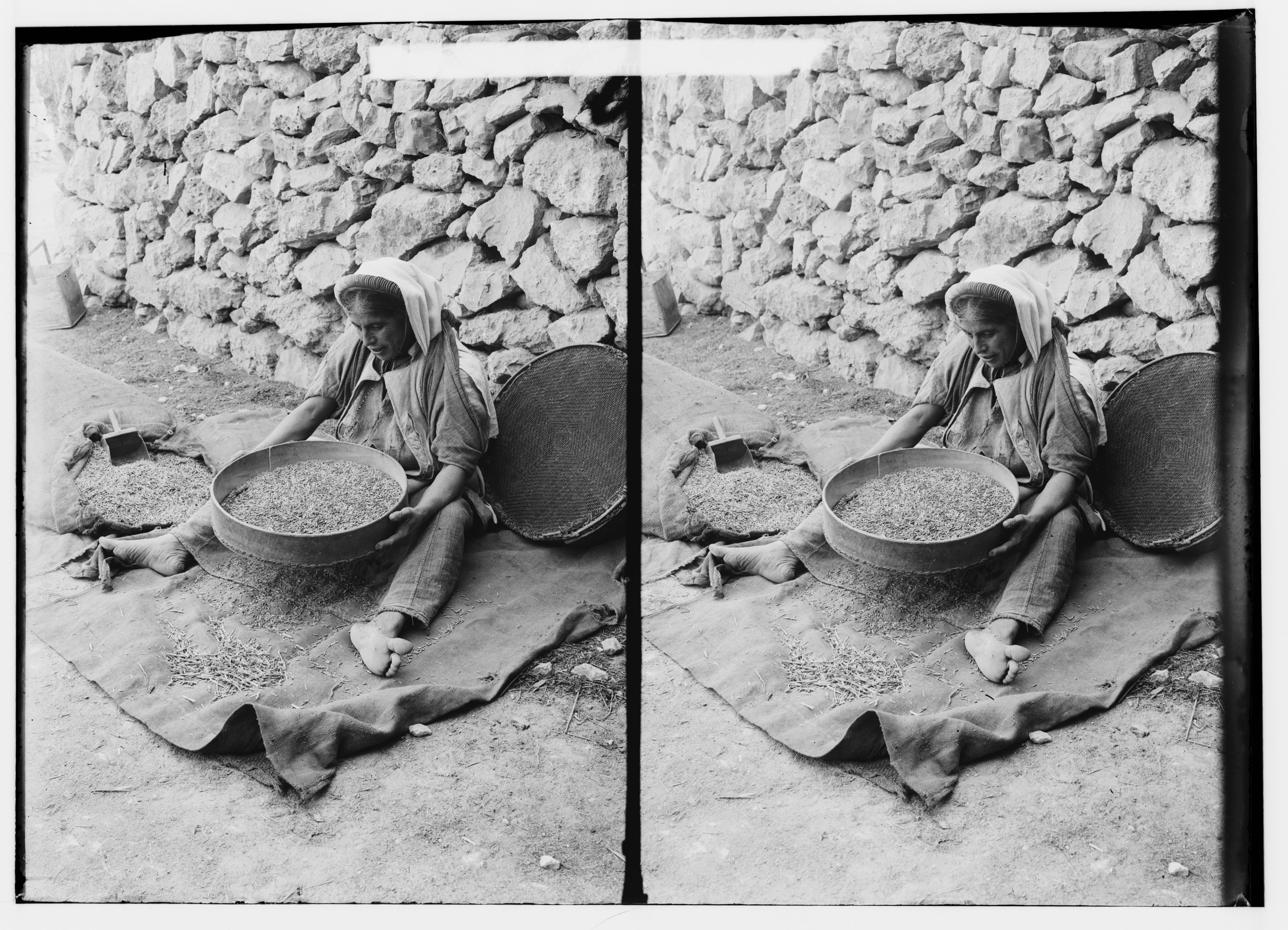 Stereoscopic photograph of a person winnowing grain in front of a stone wall.
