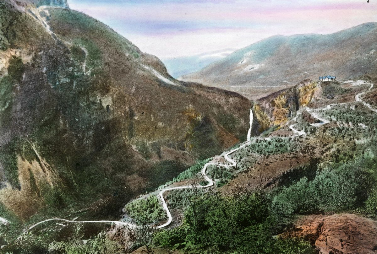Vintage hand-colored photograph of a winding road through a mountainous landscape.