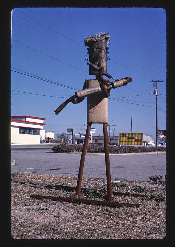 A tall, rusty metal sculpture stands in a grassy area next to a road, with buildings and a clear blue sky in the background.