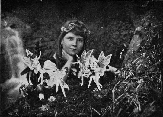 A vintage black and white photograph of a person surrounded by miniature fairy figures in a dense foliage setting.