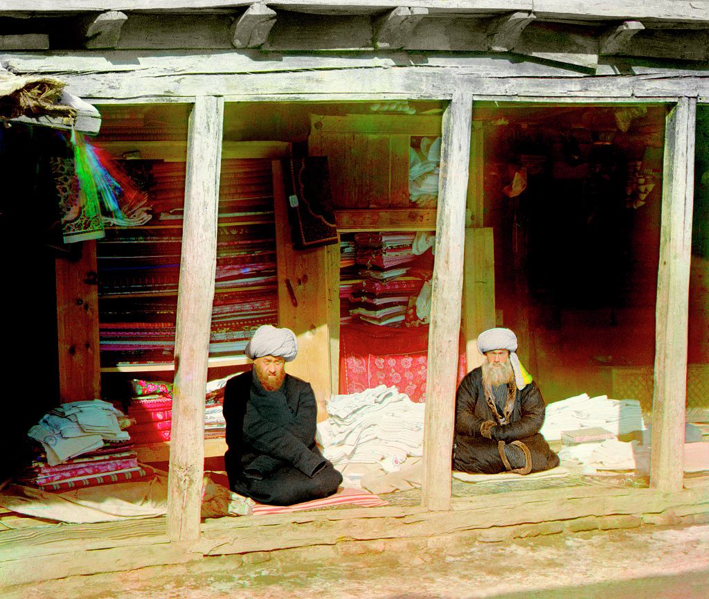 Two elderly men sit inside a wooden structure surrounded by stacks of colorful textiles.