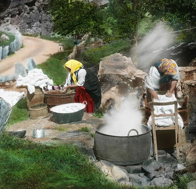 Vintage color photograph of two women doing laundry outdoors, with a large pot of steaming water and piles of white clothes.