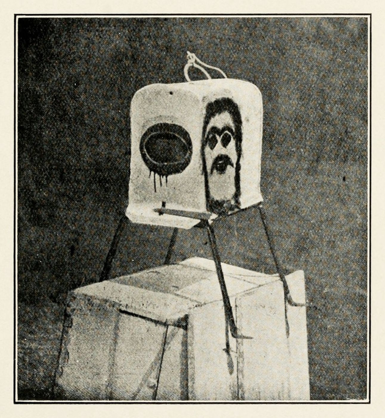 A grainy black and white photograph of a box-like structure with a face painted on it.