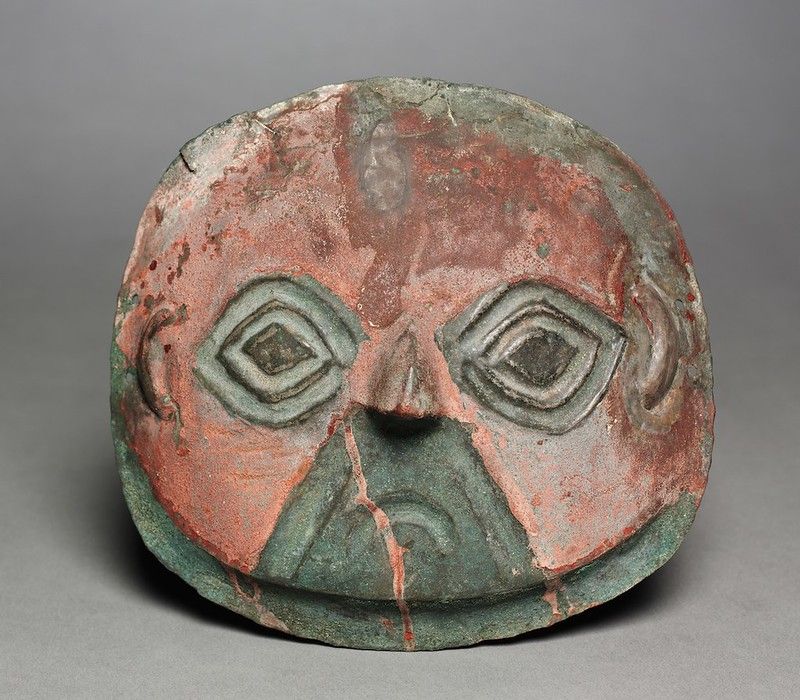 A weathered, ancient-looking mask with diamond-shaped eyes and a narrow slit mouth.