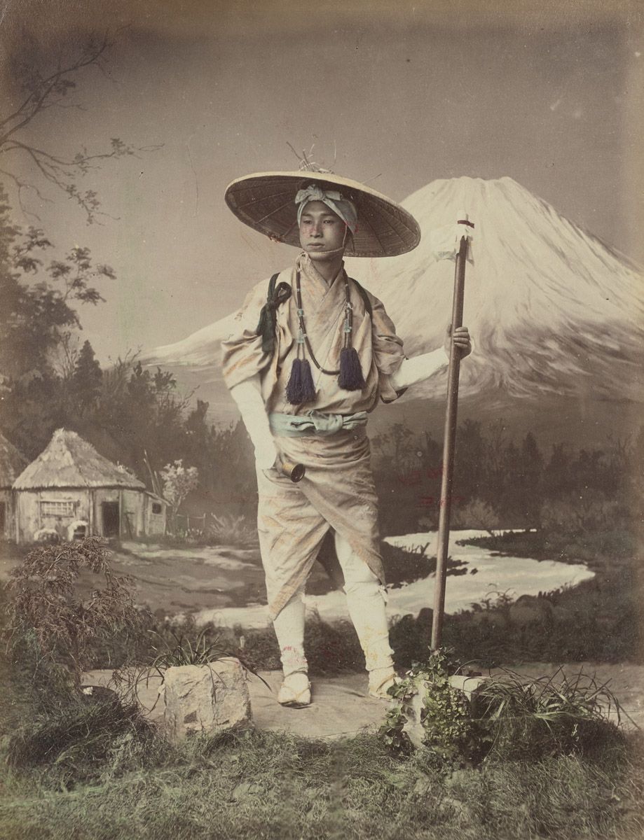 Sepia-toned photograph of a Japanese man in traditional clothing standing in front of a painted backdrop of Mount Fuji.