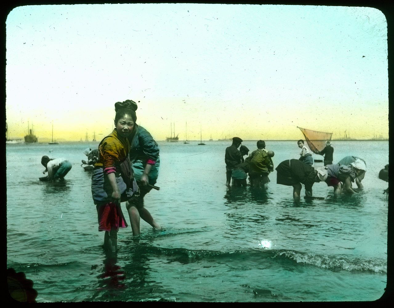 A vintage color photograph of people wading in shallow water, collecting shellfish or seaweed, with boats on the horizon and a soft gradient sky.