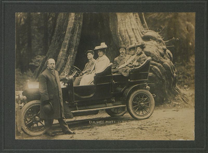 A sepia-toned vintage photograph of a group of people posing with an early automobile in a forest setting.