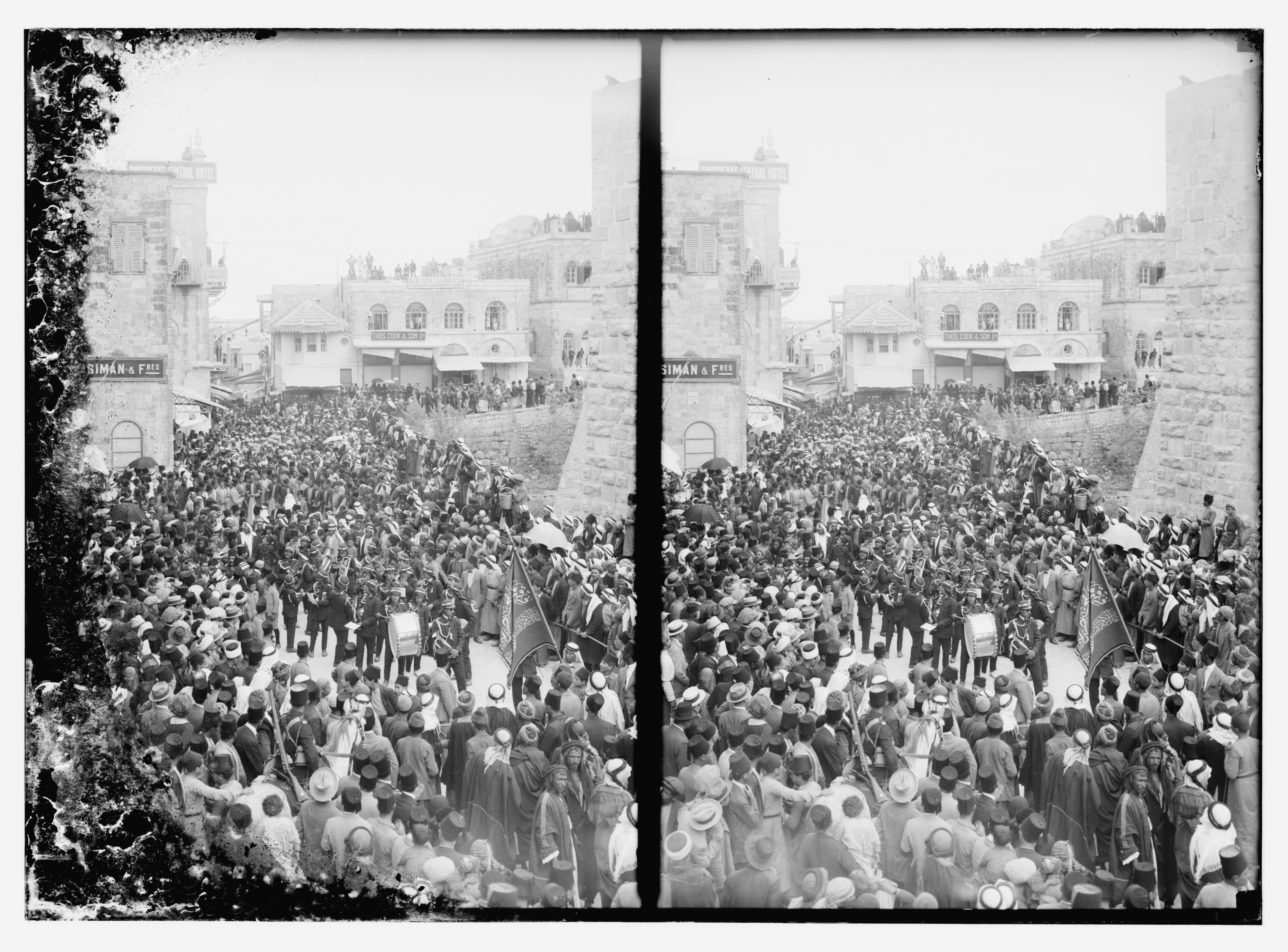 Stereoscopic photograph of a large crowd gathered in a street or square, facing a building with arched entrances and ornate details. A horse-drawn carriage or a group of people on horseback is in the center of the crowd.