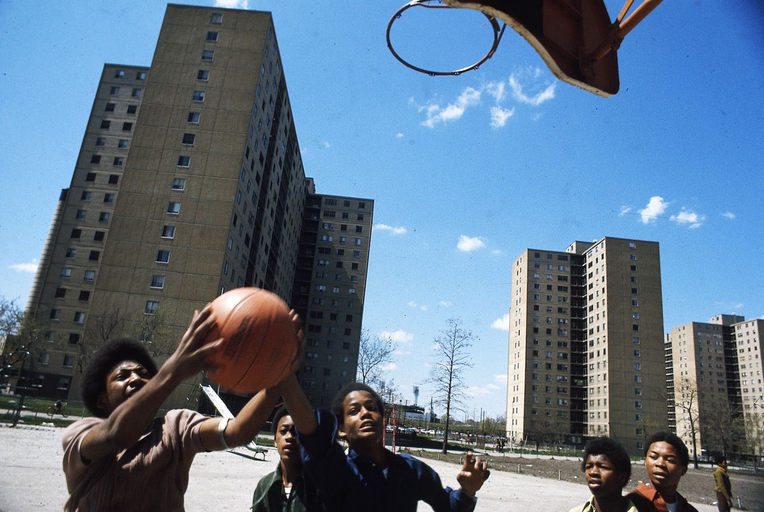 A low-angle shot of a group of young African American boys playing basketball in front of towering high-rise buildings on a sunny day.