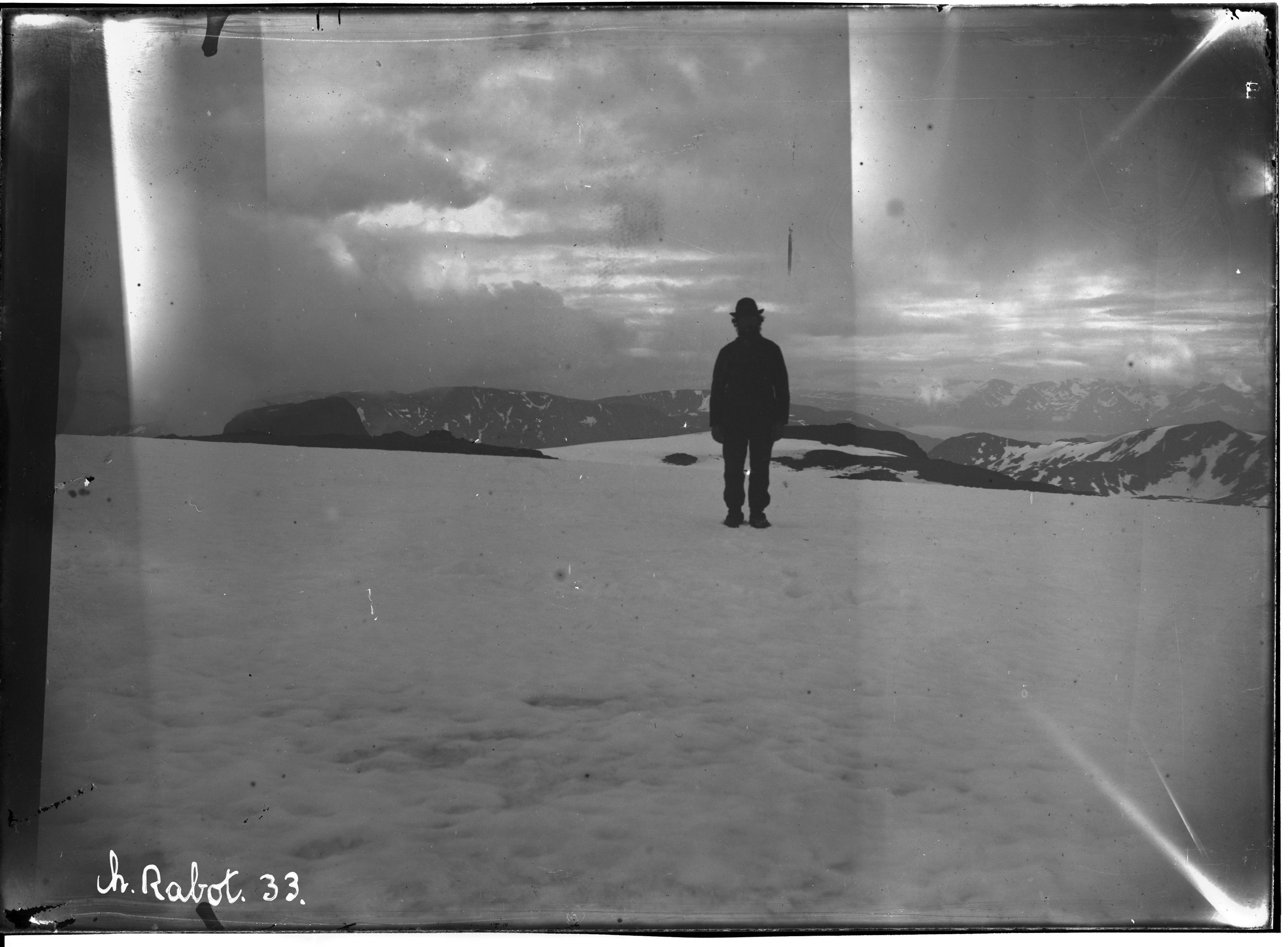 A vintage black and white photograph of a person standing in a snowy landscape with mountains in the background.