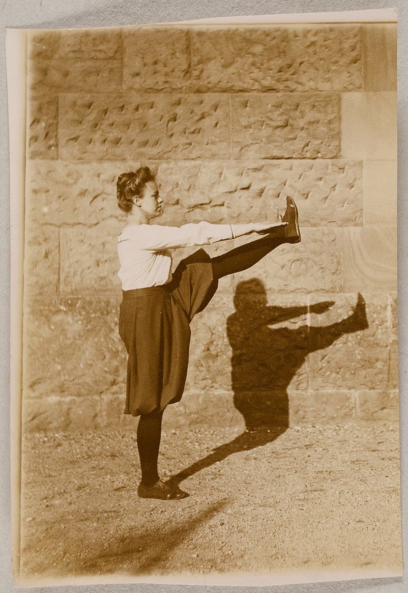 Sepia-toned photograph of a young woman performing a leg extension exercise in front of a stone wall.