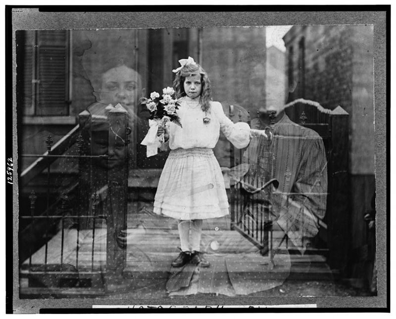 A black and white photograph of a young girl in a dress and hat holding flowers, standing on a porch or stairs.