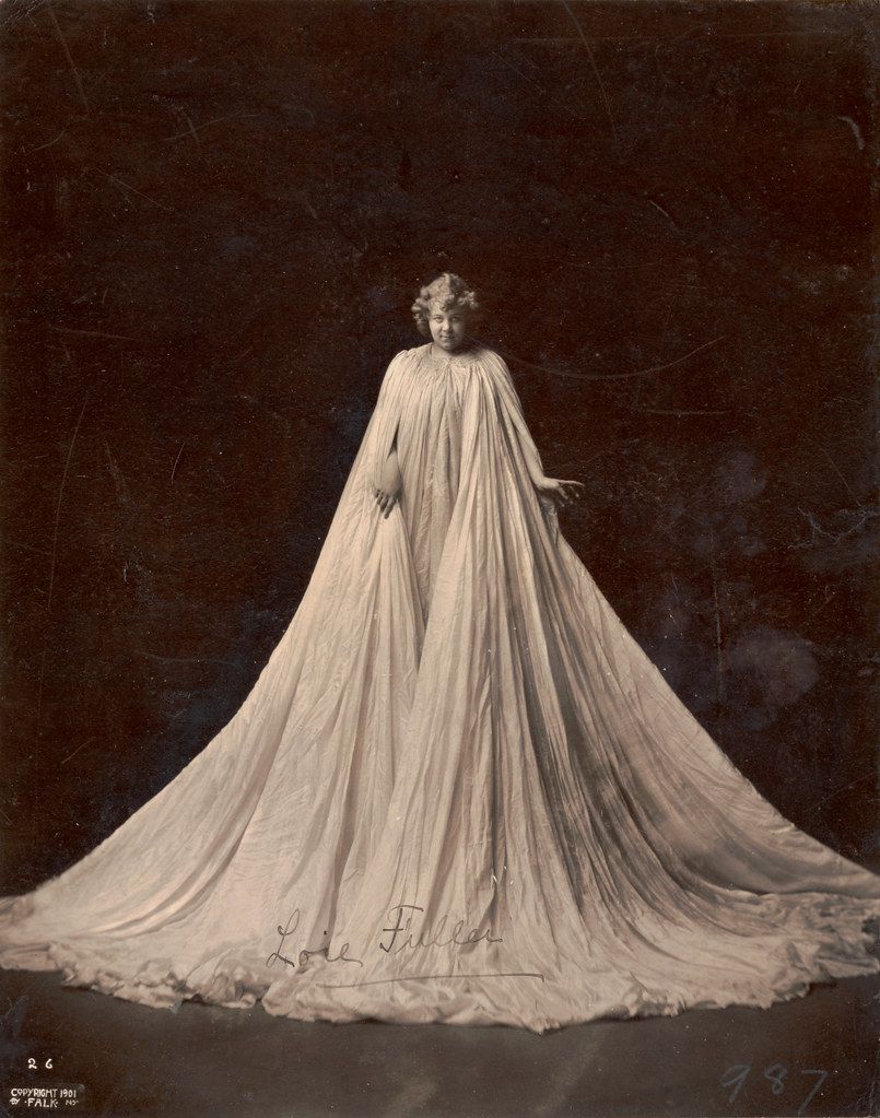 A vintage photograph of a woman in a flowing, elegant dress with a long train and cape.