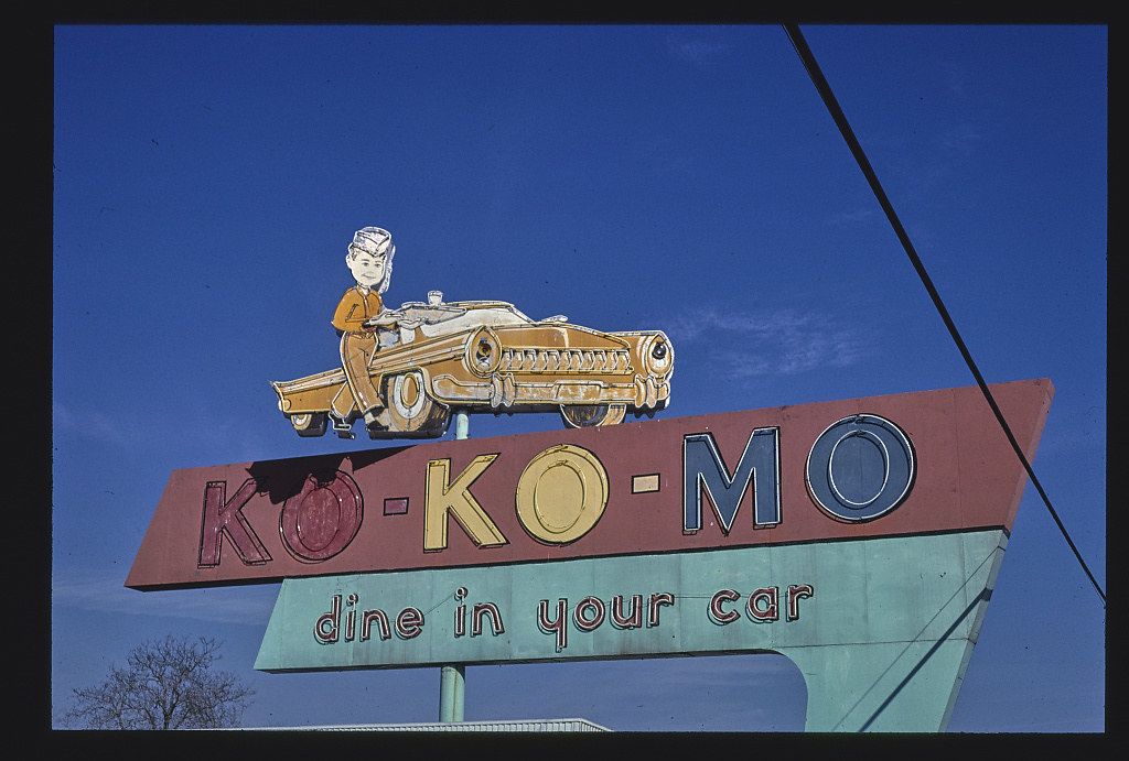 A vintage sign for the Ko-Ko-Mo drive-in restaurant, featuring a cartoon man driving a car above the restaurant's name.